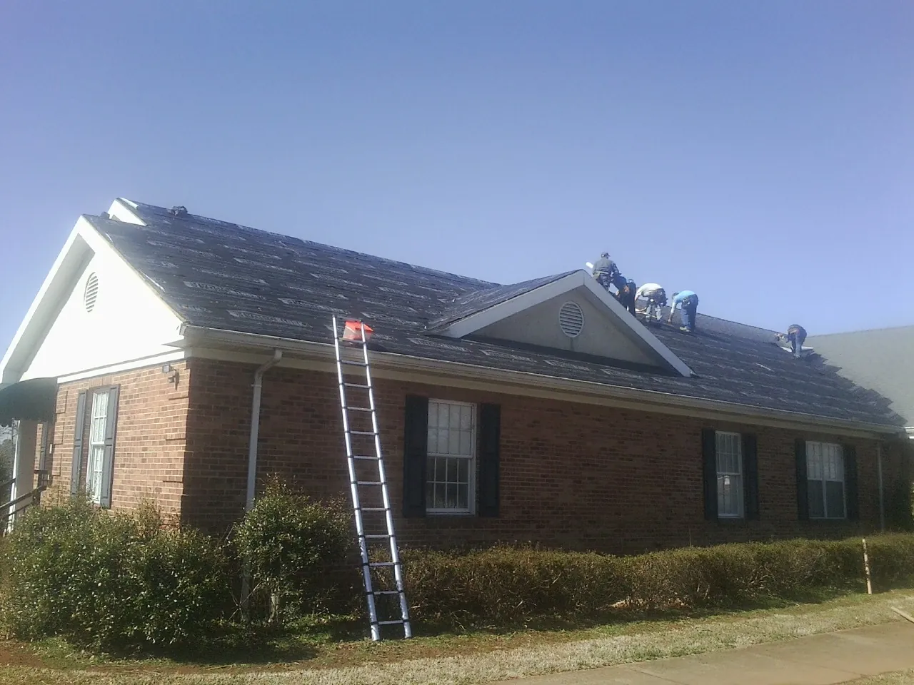 Crew tearing shingles off a brick ranch roof with a ladder set — Metal Roof Repair work by our crew in Albertville