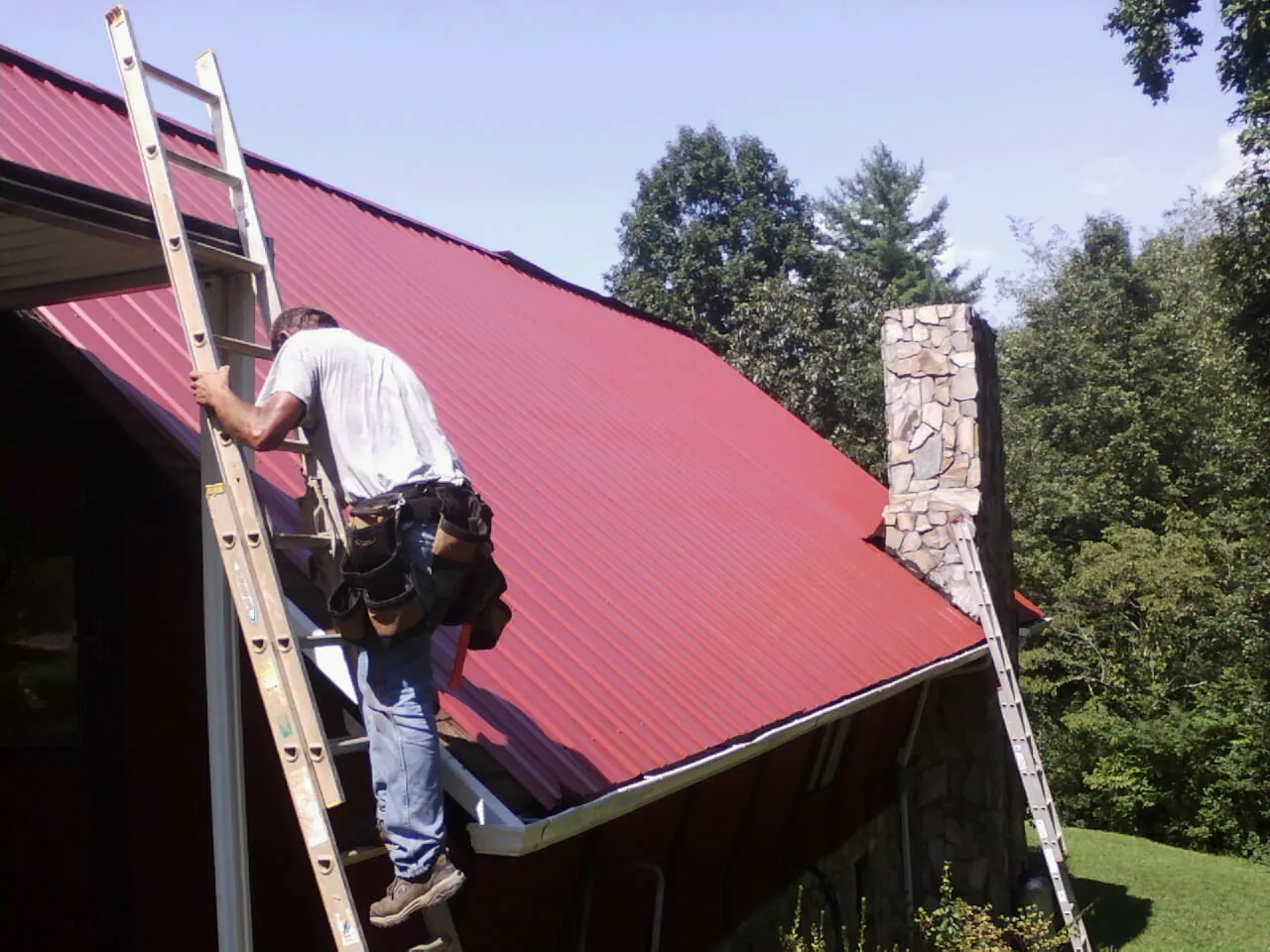 Roofer on a ladder beside a red metal roof and stone chimney — Soffit & Fascia Repair by Roofing Brothers in Albertville